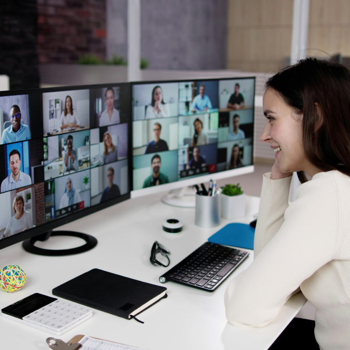 Woman smiling during an online meeting with multiple participants displayed on dual monitors, representing engaging live webinar experiences.